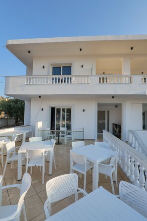 A two-story building with a balcony, outdoor seating area with white tables and chairs, and a clear sky. - Vila Eduart (Ksamil)