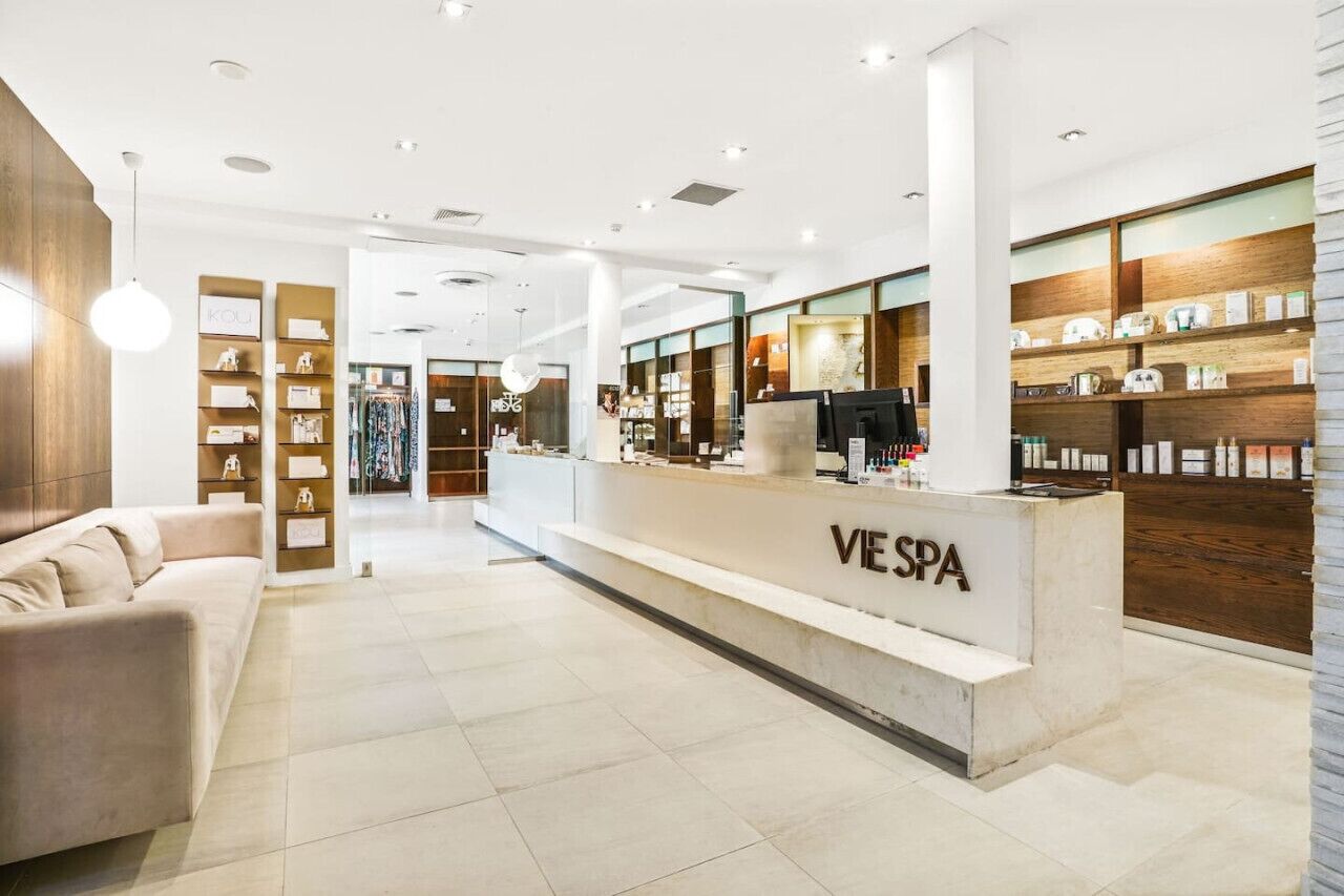 A modern spa reception area with a reception desk labeled 'VESPA,' a seating area with a sofa, and shelves with various items.