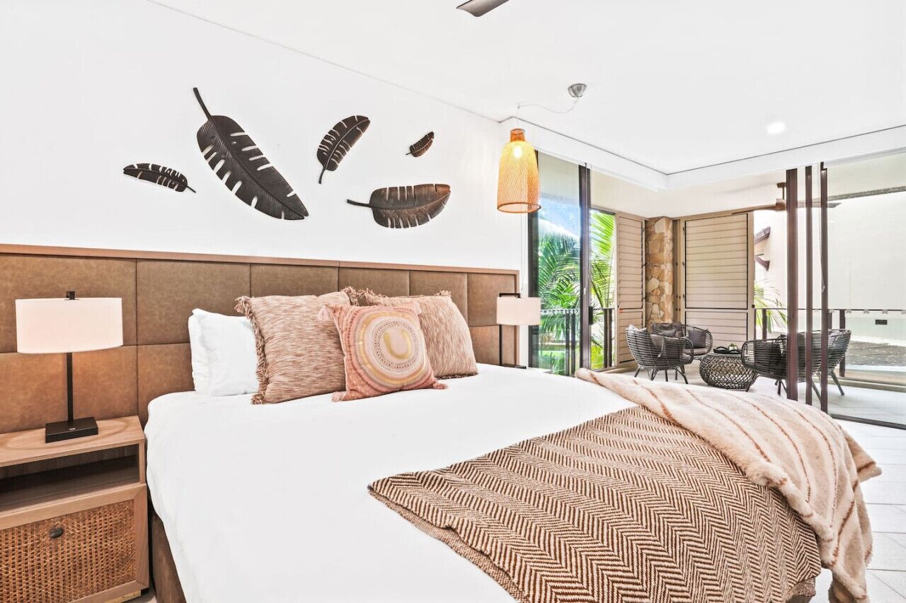 A modern hotel room with a large bed, a nightstand, a lamp, a wall-mounted artwork, a hanging light fixture, and a balcony with outdoor seating.