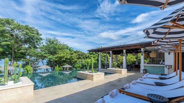 Zona junto a la piscina con sillones, un bar y vista al mar.