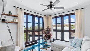 Amplia sala de estar con un sofá blanco, una mesa de centro de vidrio y un ventilador de techo. Amplias ventanas ofrecen vistas al exterior.