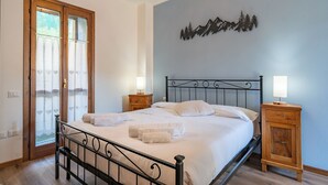 Een net opgemaakt bed met witte lakens en handdoeken, een houten nachtkastje met een lamp, en een kunstwerk aan de muur met daarop een berglandschap.
