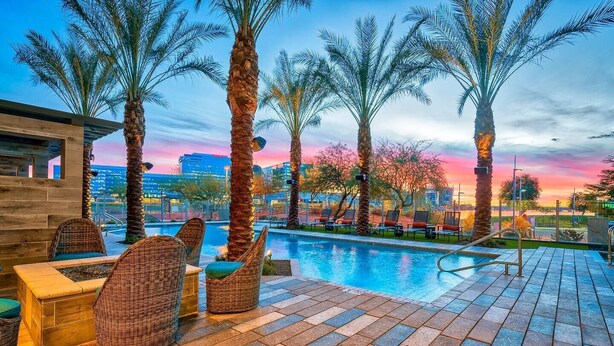Zona junto a la piscina con mobiliario de mimbre y palmeras, con vistas a un paisaje urbano al atardecer.