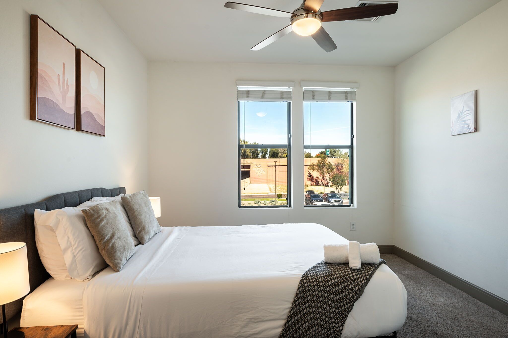A hotel room with a bed, a ceiling fan, a window with a view, and artwork on the walls.