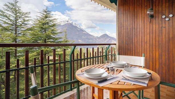 A wooden balcony with a table set for two, overlooking a mountainous landscape with trees and a clear sky.