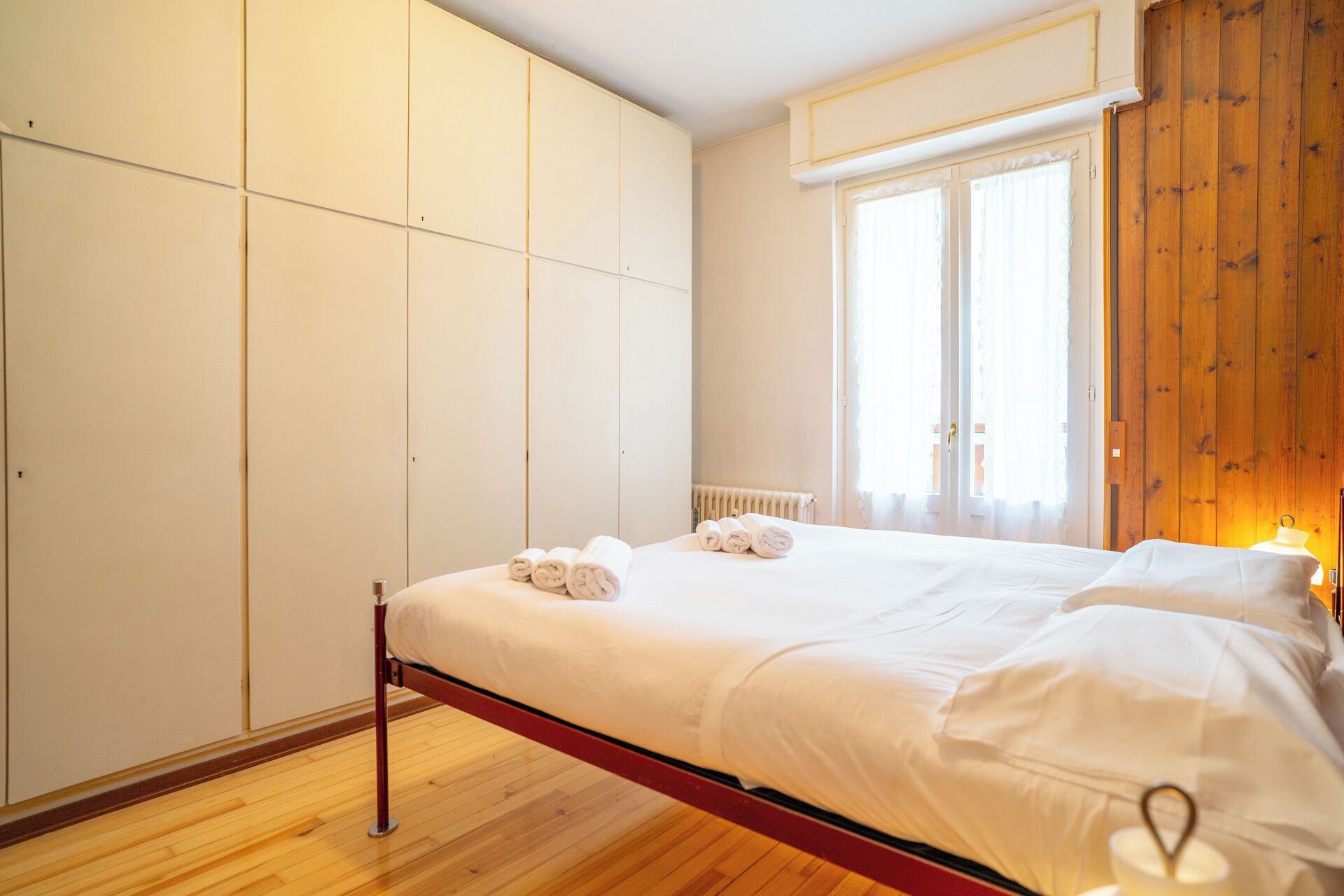 A bedroom with a bed, wooden floor, white walls, and a window with curtains.