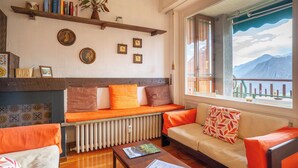 A cozy room with a sofa, a bench, and a table. There are books and a vase on a shelf. A window offers a view of mountains.
