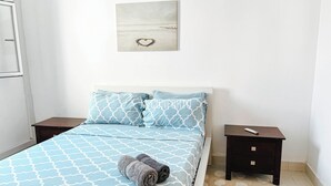 A neatly made bed with a patterned bedspread, two pillows, and a rolled towel. There is a small bedside table with a phone and a wall-mounted picture. - Remarkable 5-bed Apartment in Kilamba - Luanda (Belas)