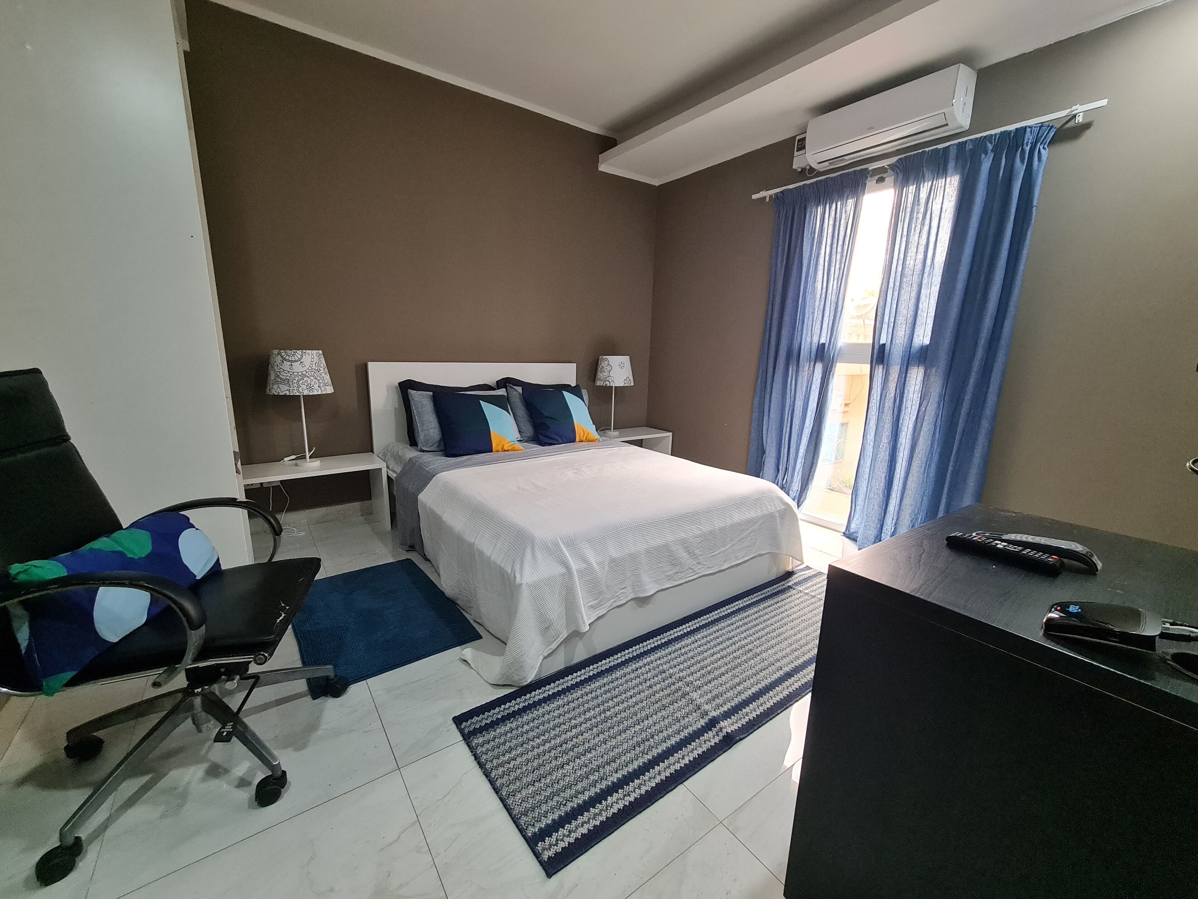 Una habitación de hotel con cama, mesitas de noche, un escritorio, una silla y un aire acondicionado.