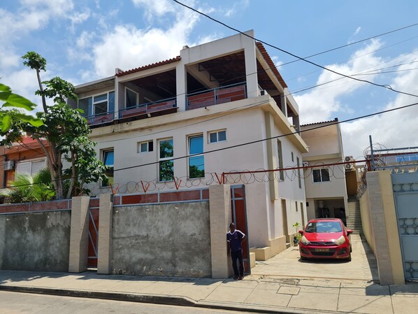 Un edificio de dos pisos con un balcón, una entrada protegida por una reja y un coche rojo estacionado al frente.