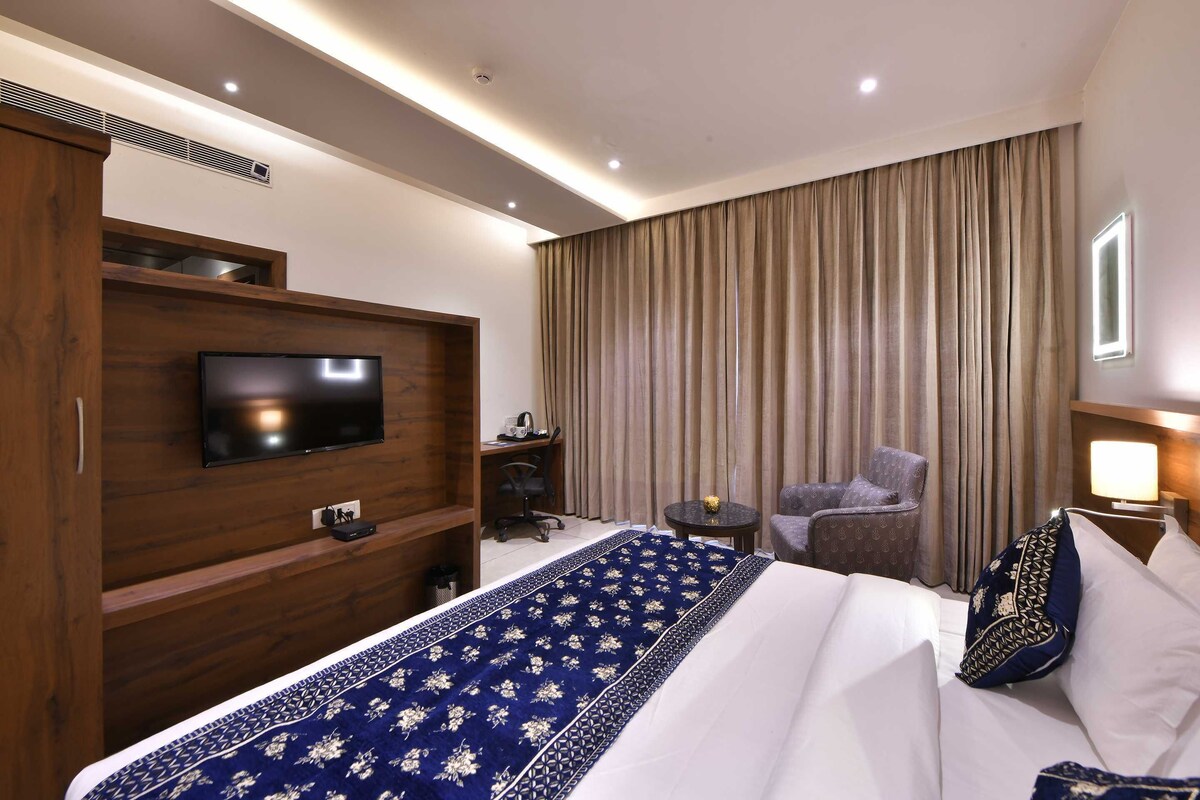 Hotelkamer met groot bed, televisie bevestigd aan een houten paneel, bureau met stoel, klein tafeltje met lamp en raam met gordijnen.