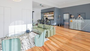 A modern lobby with a reception desk, a green sofa, a glass table, and a wooden floor. - Punthill Sunshine (Sunshine)