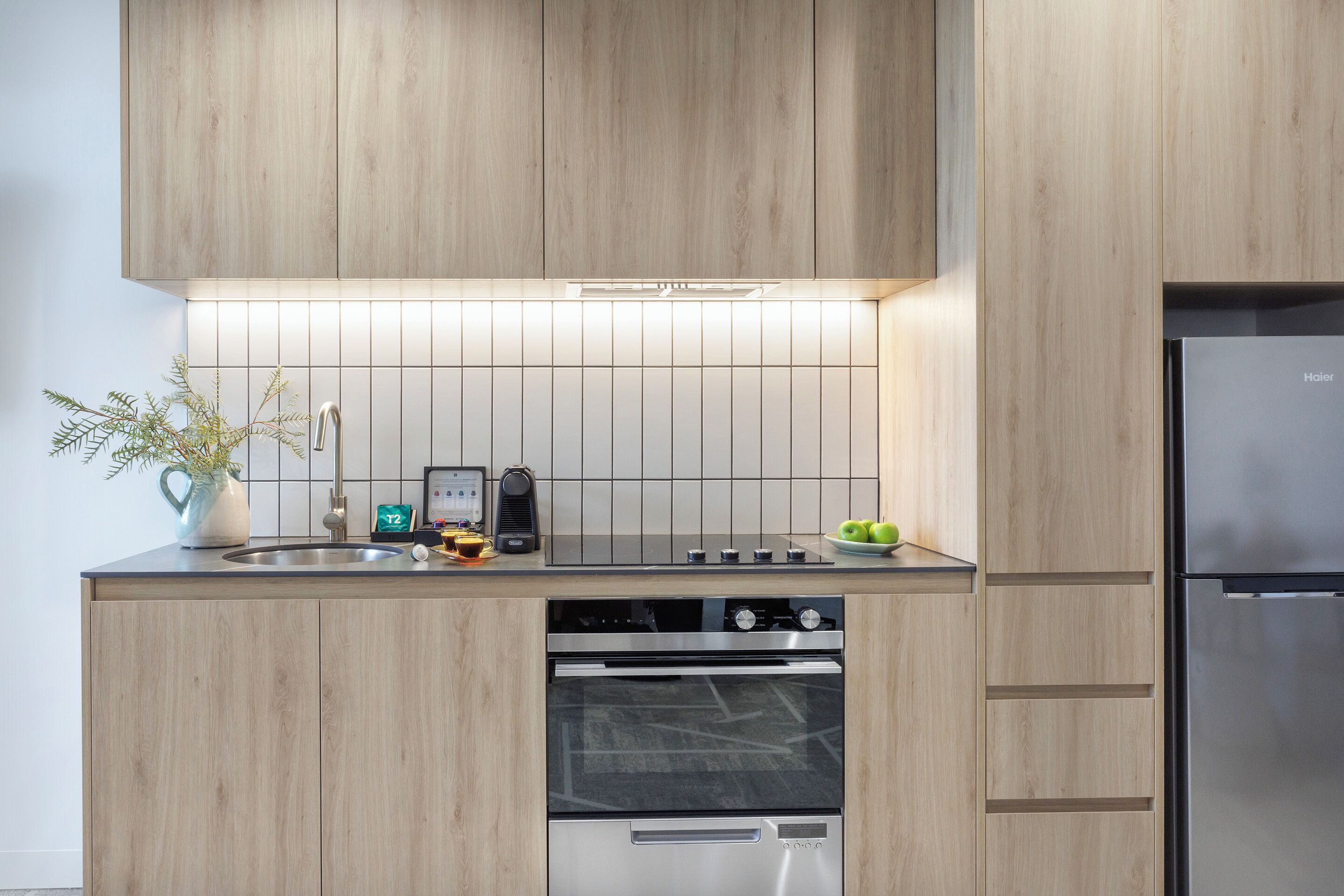A modern kitchen with wooden cabinets, a stainless steel refrigerator, a built-in oven, and a tiled backsplash.