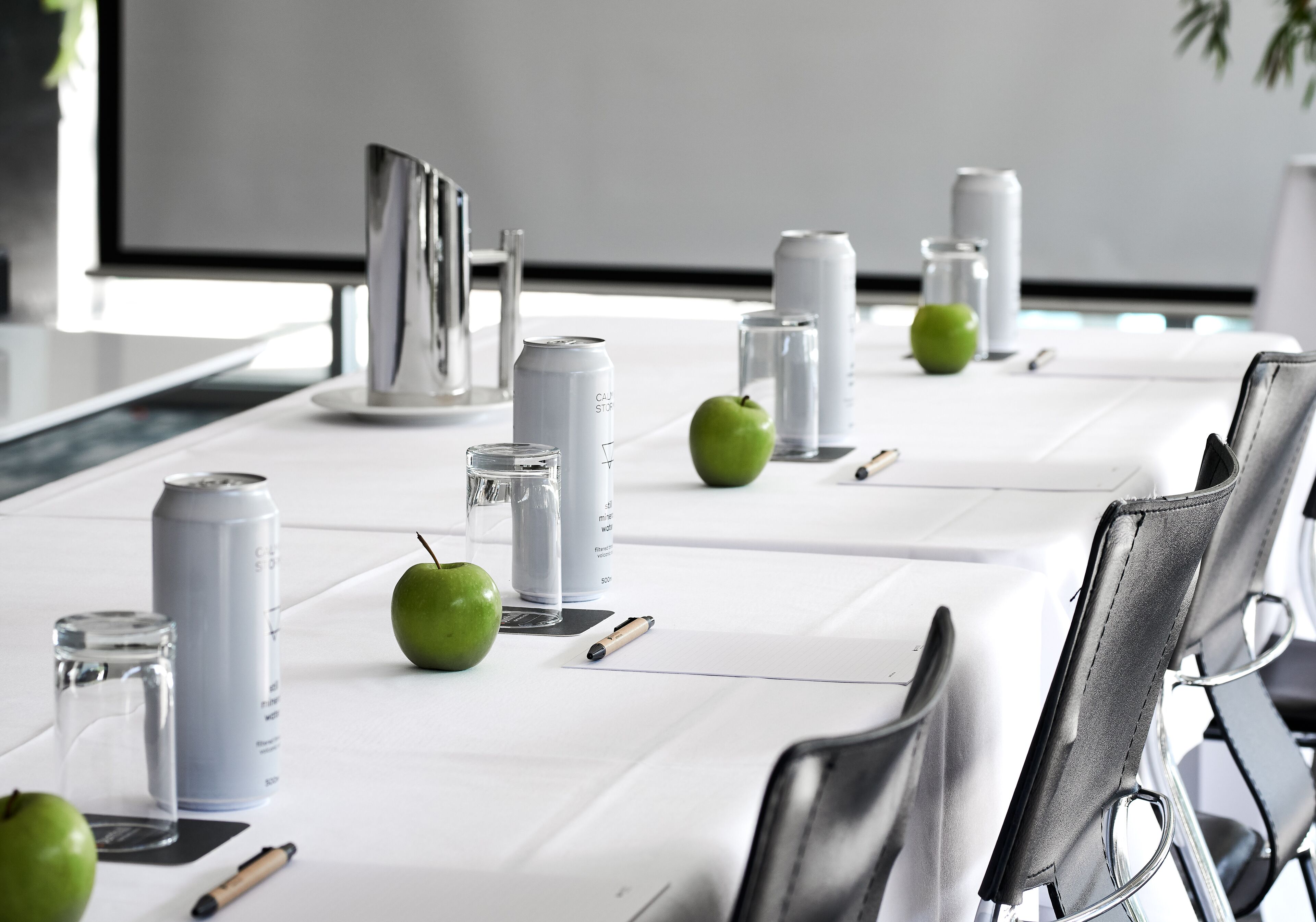 A conference table with white tablecloth, silver canisters, green apples, and a glass of water.