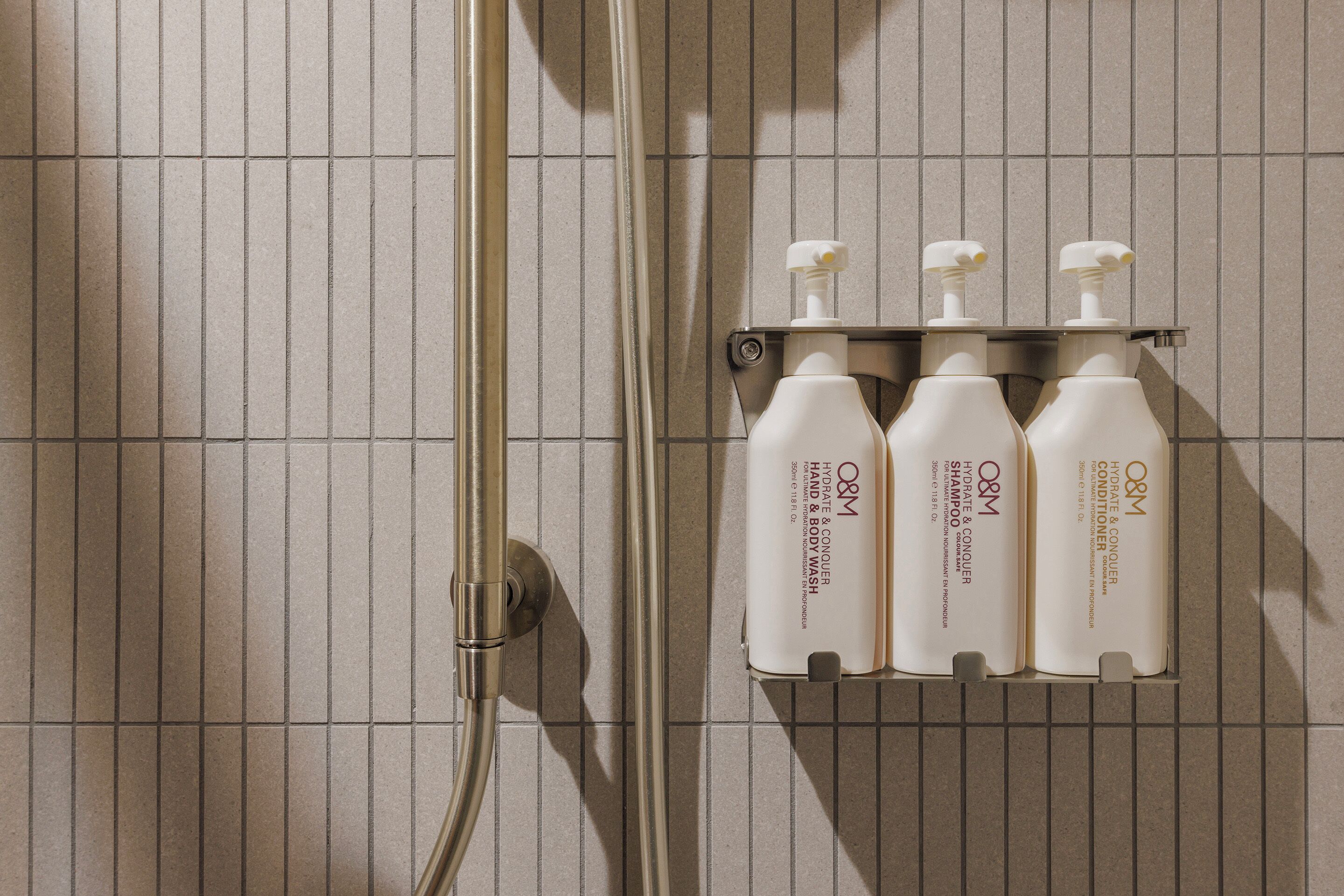 Un baño con pared de azulejos, una ducha y un estante con cuatro botellas de jabón de manos y cuerpo O&M.