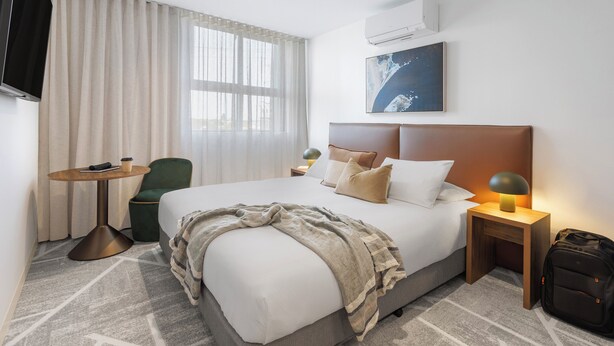 A hotel room with a large bed, a bedside table with a lamp, a chair, a TV, and a window with curtains. - Punthill Sunshine (Sunshine)