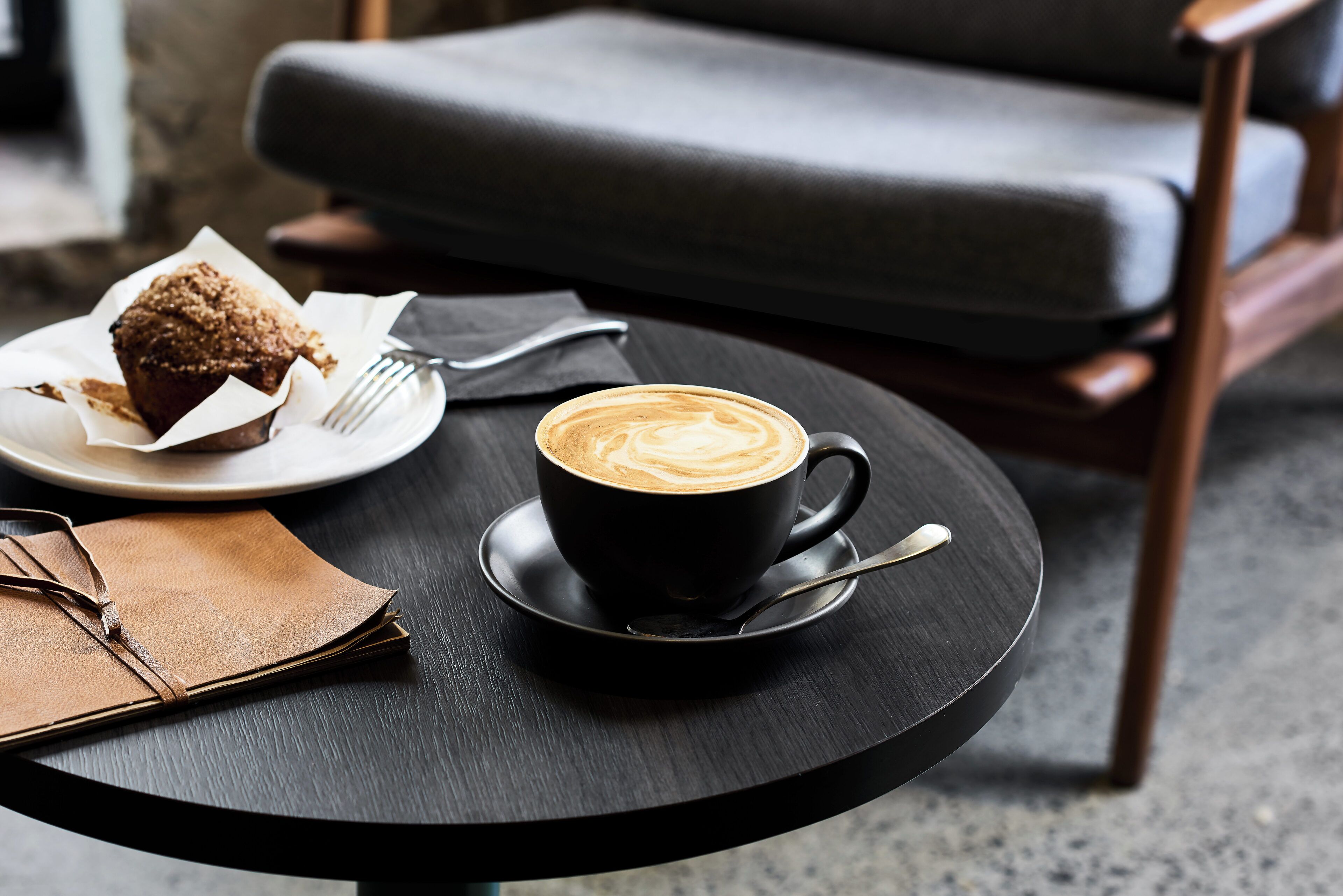 A black cup of coffee with latte art on a black saucer, a muffin on a white plate, a brown leather bag, and a black chair in the background.