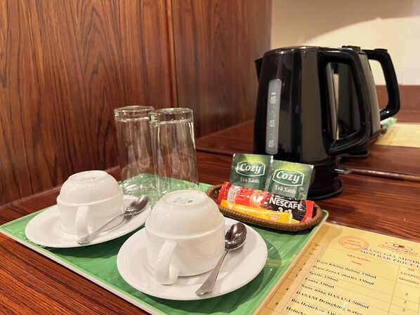 Une table en bois est dressée pour le petit-déjeuner avec deux tasses blanches, deux cuillères, deux verres transparents, deux sachets de thé « Cozy », un sachet de café instantané « Nescafe » et une cafetière noire.