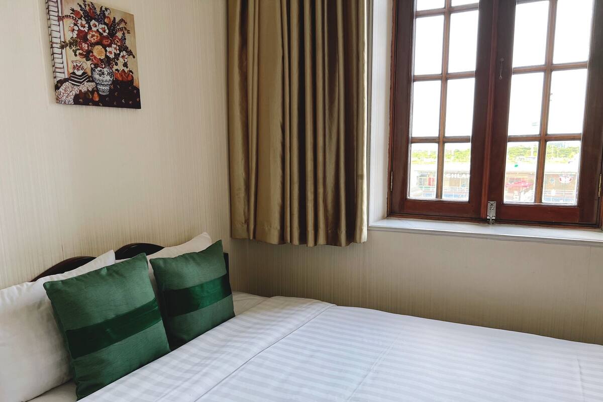 A hotel room with a bed, green pillows, a painting on the wall, and a window with curtains.
