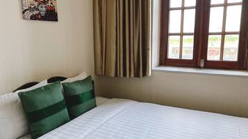 A hotel room with a bed, green pillows, a painting on the wall, and a window with curtains.