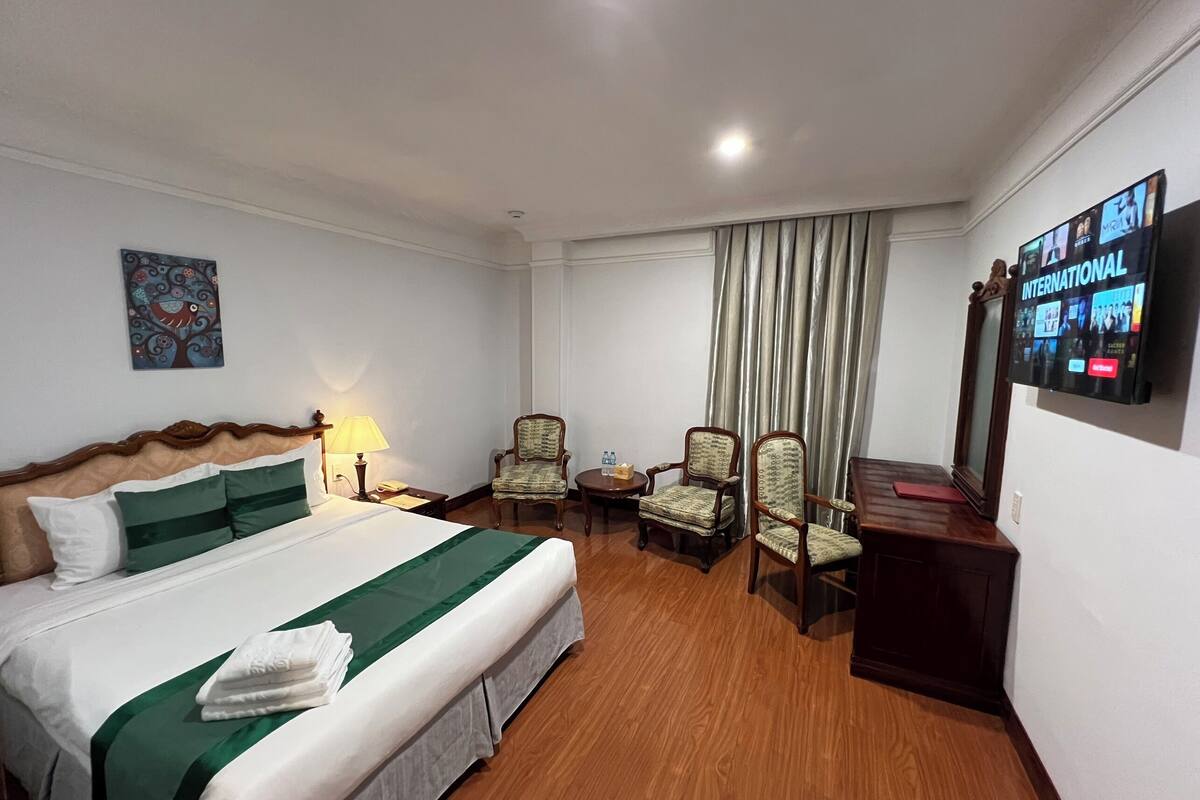 A hotel room with a bed, two armchairs, a small table, a TV, and a painting on the wall.