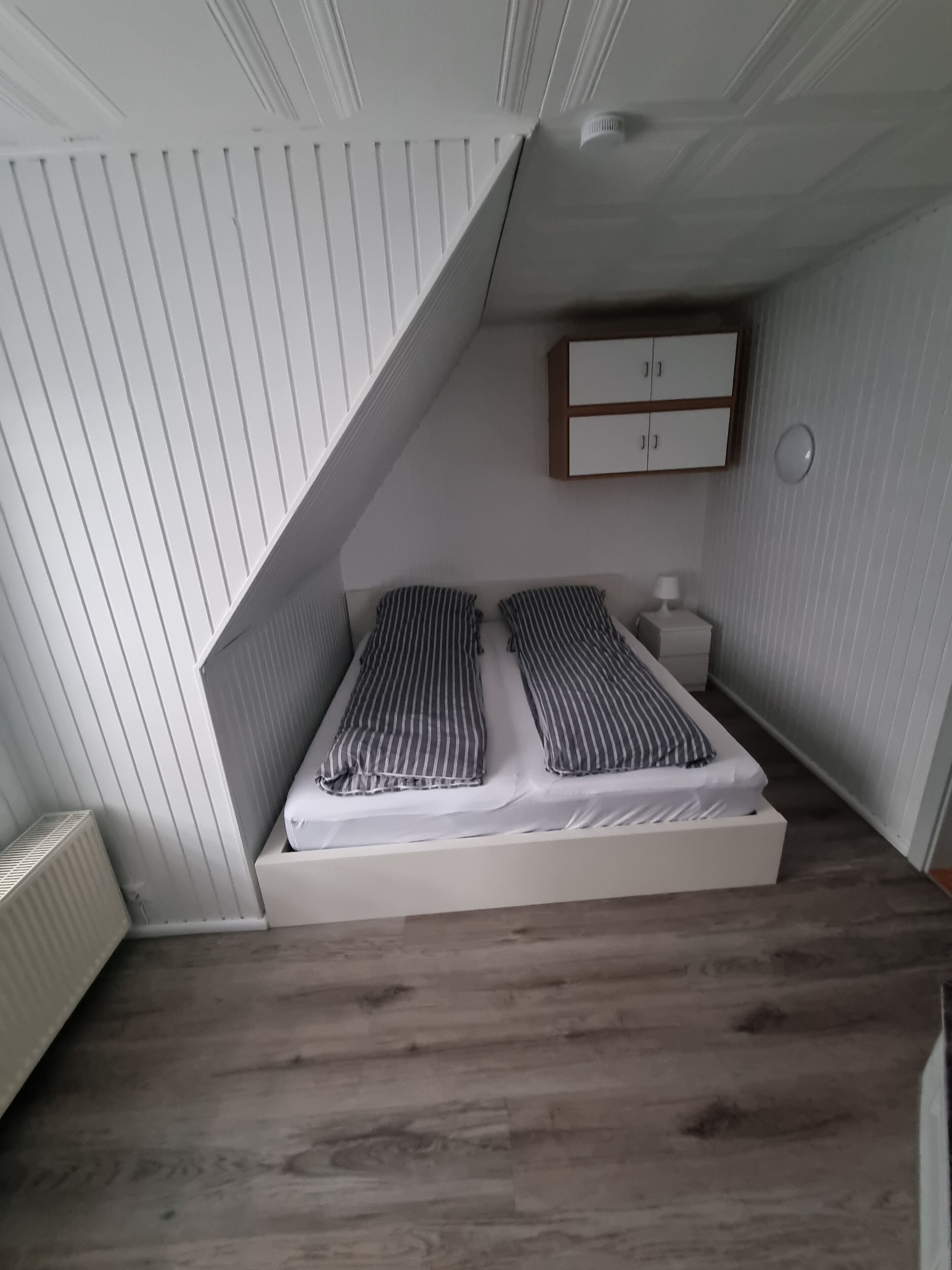Quarto pequeno com teto inclinado, uma cama de solteiro, uma mesa de cabeceira e um armário.
