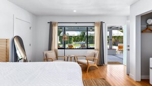 Ein modernes Hotelzimmer mit einem großen Bett, zwei Sesseln, einem kleinen Tisch und Blick ins Freie durch ein großes Fenster.