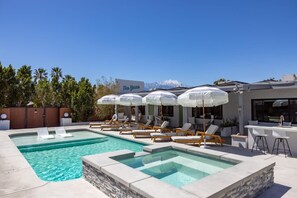 Zona de piscina con sillas de descanso y sombrillas, un bar y vistas a la montaña.