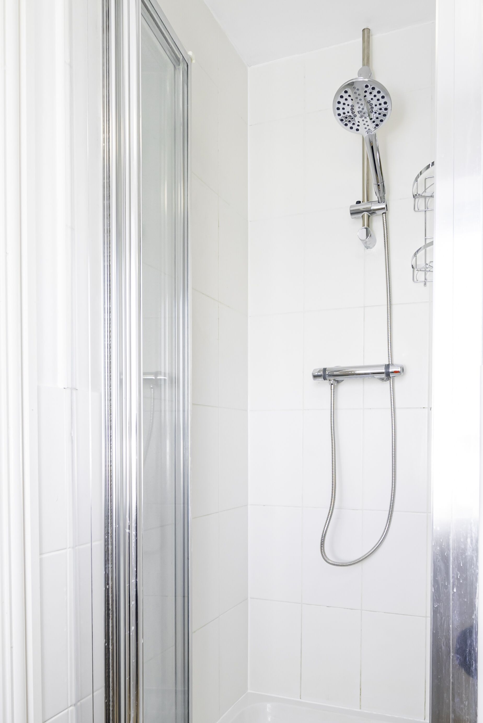 Une douche moderne équipée d’un pommeau de douche à main et d’un pommeau de douche fixe fixé au mur, tous deux dotés de finitions chromées, dans une salle de bain aux murs recouverts de carreaux blancs.