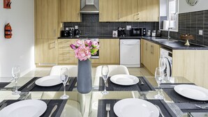 A modern kitchen with wooden cabinets, a black tiled backsplash, and a dining area set with a glass table, white chairs, and a vase with flowers. - Modern Home, 9 beds for Extended Stays, Free Parking (Reading)
