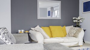 A modern living room with a grey sofa, a white sofa with yellow cushions, a small side table with a tray of croissants, and a mirror on the wall. - Modern Home, 9 beds for Extended Stays, Free Parking (Reading)