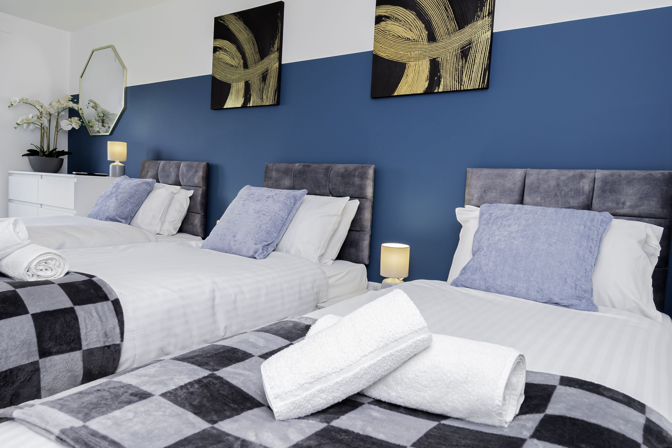 A hotel room with two beds, each with a grey headboard and white bedding. There are two wall-mounted paintings, a bedside table with a lamp, and a potted plant.