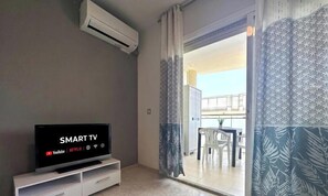A room with a flat-screen TV displaying 'SMART TV' and streaming services logos, a white entertainment unit, a sliding glass door leading to a balcony with a dining table and chairs.