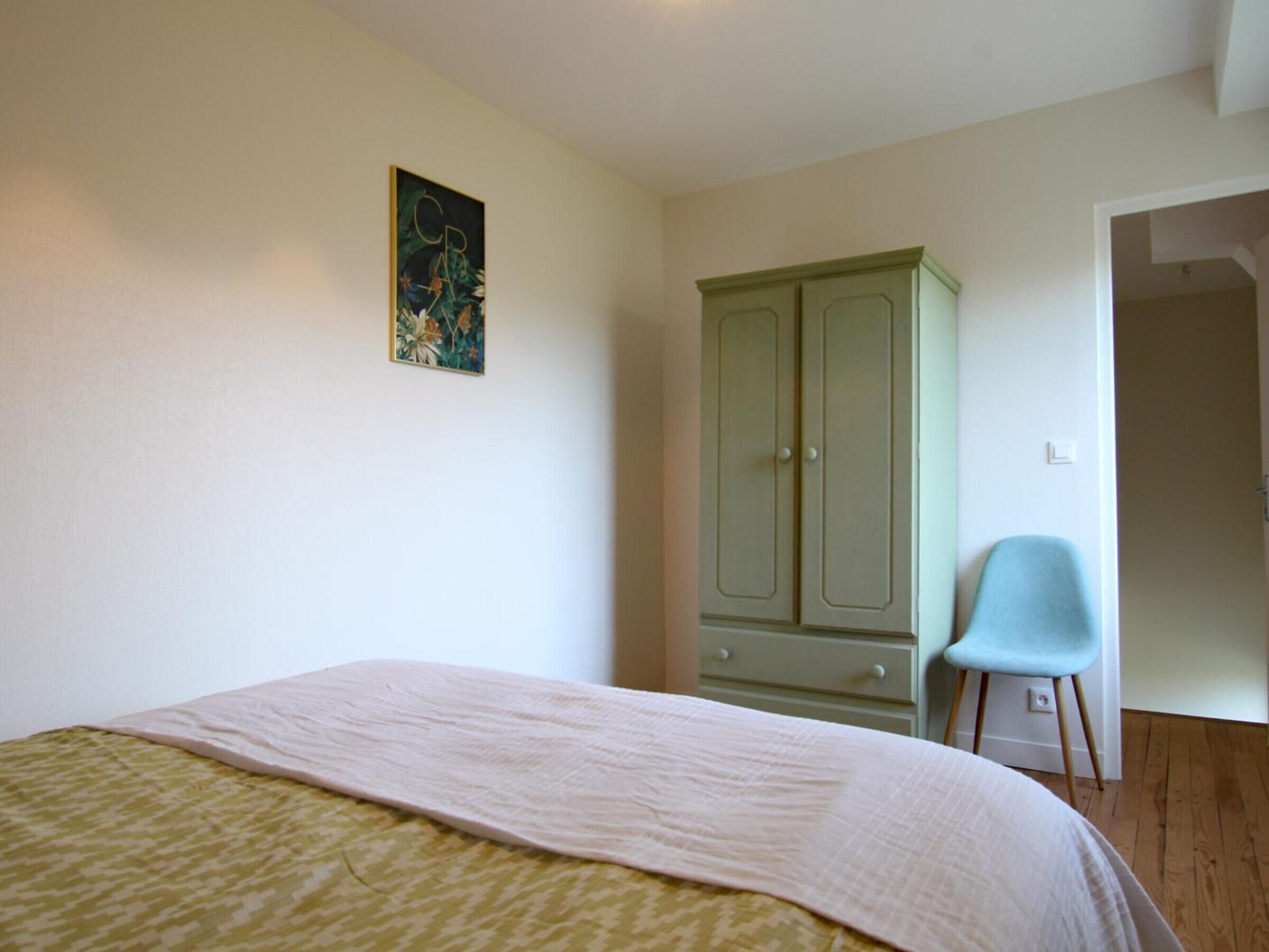 Un dormitori amb un llit, un armari verd, una cadira i una porta que dona a una altra habitació.