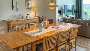 A dining area with a wooden table and chairs, a vase with flowers, a lamp, and a television in the background.
