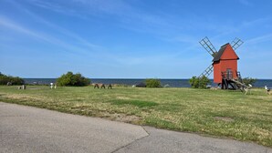 A red windmill with a wooden structure and a metal roof, situated on a grassy field with a paved path in the foreground.