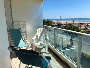 Ein Balkon mit zwei grünen Loungestühlen und einem weißen Stuhl mit Blick auf einen Strand, auf dem Autos geparkt sind, und einem wolkenlosen Himmel.