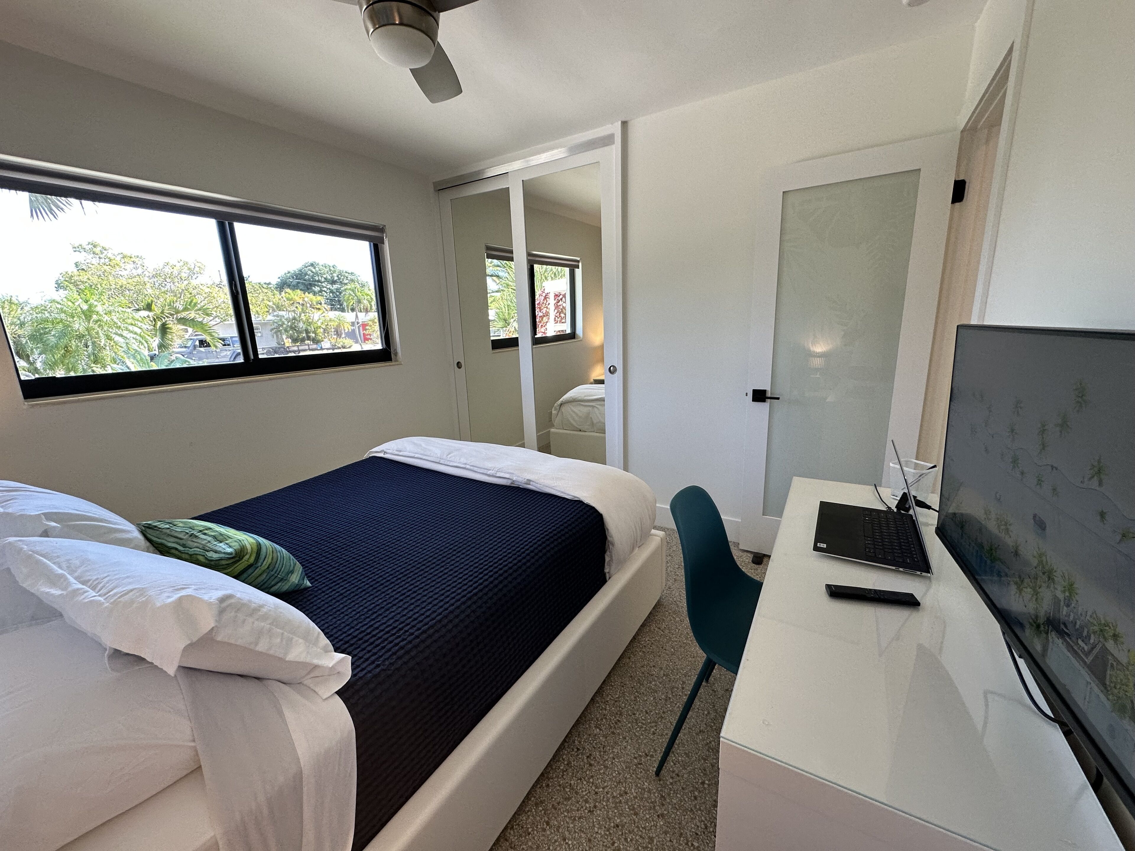 Une chambre à coucher avec un lit, un bureau et un ordinateur. Il y a une fenêtre donnant sur des arbres et une porte qui mène à une autre pièce.