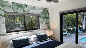 A bedroom with a large bed, a window with a view of a pool and greenery, a ceiling fan, and a nightstand with a lamp.