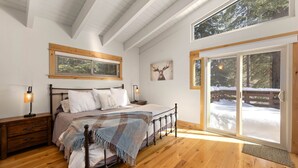 Een slaapkamer met een houten bed, een nachtkastje, een raam met blinds, een deur naar buiten en een aan de muur bevestigd kunstwerk van een hertenkop.
