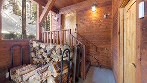 Interieur van een houten hutje met een trap, een stapel hout en een deur die naar buiten leidt.