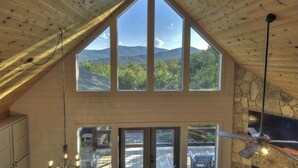 Una stanza con soffitto alto, ampie finestre che si affacciano su una montagna, un muro in pietra e una ventola a soffitto.