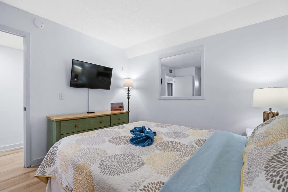 Un dormitorio con cama, televisor montado en la pared, mesita de noche con lámpara y espejo en la pared.