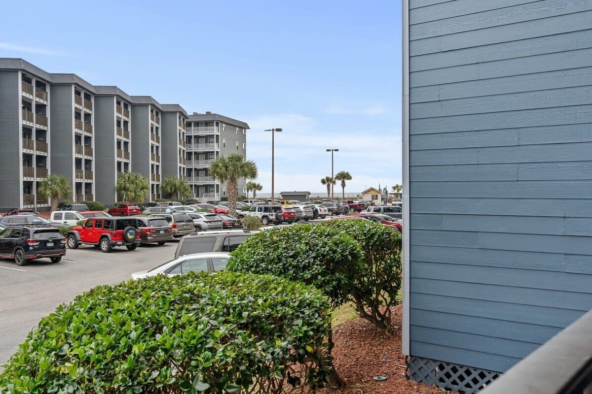A parking lot with multiple cars, a building with balconies, and a small hedge in the foreground.