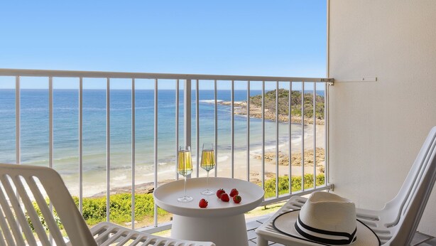 Un balcon donnant sur la plage, une table avec deux verres et des fraises, et un chapeau posé sur une chaise.