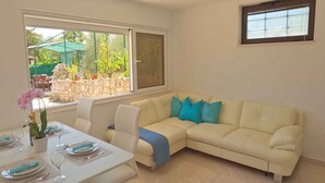 Una sala de estar con un sofá color beige, una mesa de comedor para cuatro personas y un ventanal con vistas a un jardín.