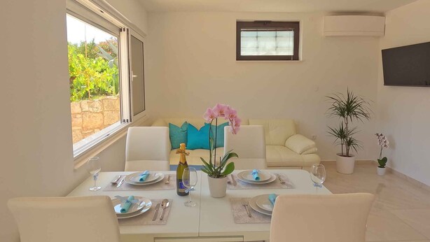 Un comedor con una mesa lista para cuatro, un sofá con cojines, una orquídea en maceta y un ventanal con vistas al verdor.