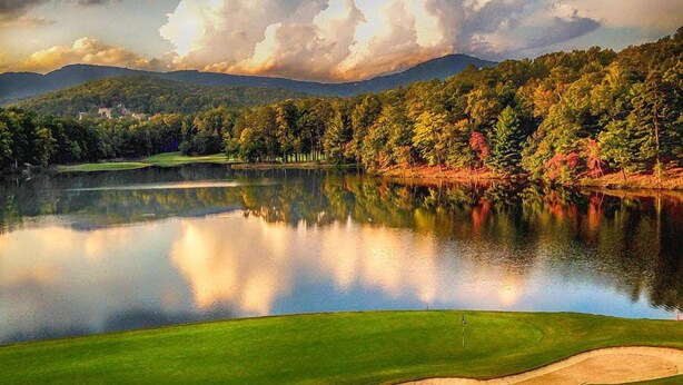 Un campo de golf tranquilo con un lago sereno, rodeado de bosques densos y montañas al fondo.
