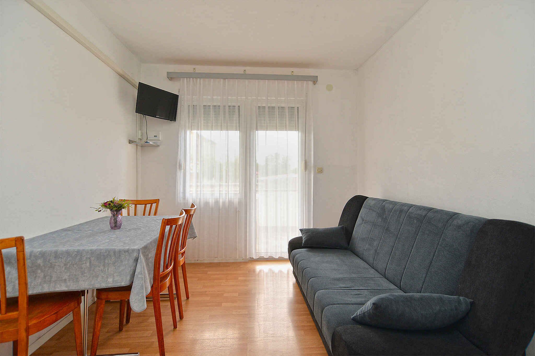 Ein Zimmer mit grauem Sofa, Esstisch aus Holz, Stühlen und einem an der Wand befestigten Fernseher.