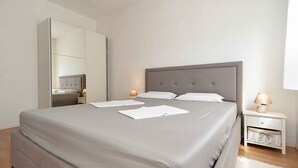 Un lit bien fait, avec une tête de lit grise, une table de chevet avec une lampe et un grand miroir accroché au mur.